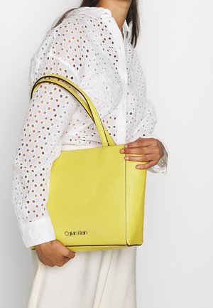 Yellow leather tote bag with a smooth texture, featuring black trim and logo accent. Worn over the shoulder with long straps.