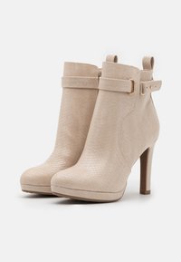 Bottines beige à talons hauts avec motif texturé, brides latérales à boucle et languettes à l’arrière, photographiées sur fond blanc.