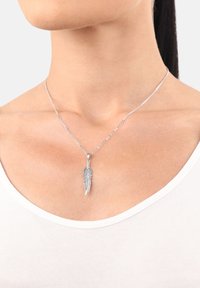 Collana con pendente a forma di piuma d'argento su una catena sottile, presenta un'incisione dettagliata delle foglie, indossata su un collo nudo sopra una maglietta bianca.