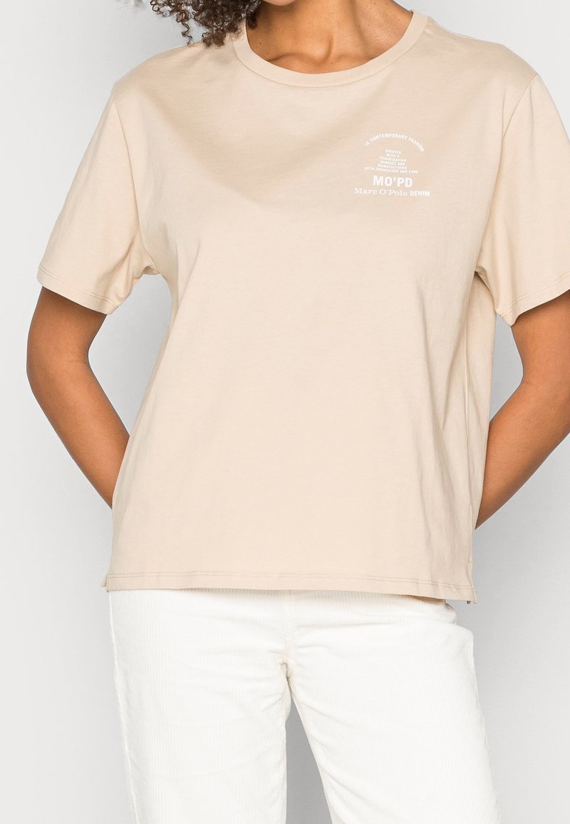 Béžové bavlněné tričko s krátkým rukávem a kulatým výstřihem, s bílým textovým grafikou na přední straně a volným střihem.