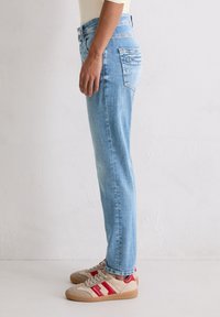 Persoon in lichtblauwe jeans en beige sneakers met rode strepen, staande in profiel tegen een effen witte muur.