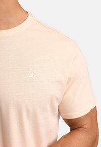 Jasno brzoskwiniowa bawełniana koszulka z okrągłym dekoltem, z małym białym logo "EST. 1993" na lewej piersi. Gładka tekstura.