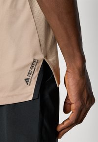 T-shirt de sport beige en tissu texturé, avec fente latérale et accents noirs. Logo "PRO-SERIES" imprimé près de l'ourlet, conçu pour améliorer la performance.