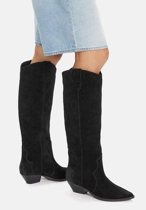 Nogi w jasnoniebieskiej dżinsowej spódnicy noszące czarne zamszowe kozaki za kolano z niskim, skośnym obcasem na białym tle.