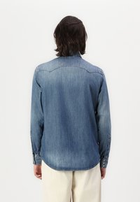 Camicia in denim blu medio con maniche lunghe, orlo curvo e polsini con bottoni. Presenta cuciture a contrasto e colletto classico.