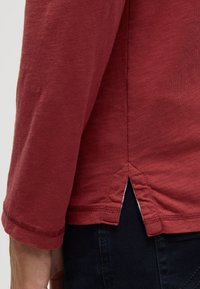 Langärmliges Hemd aus einem schlichten roten Stoff mit strukturierter Oberfläche. Verfügt über einen Seitenschlitz am Saum und kontrastierende Nähte entlang der Kanten.