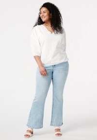 Weißes Blusenshirt mit Spitzenbesatz und drei Viertelärmeln, kombiniert mit hellblauen Schlagjeans. Das Modell trägt cremefarbene Sandalen und schlichten Schmuck.