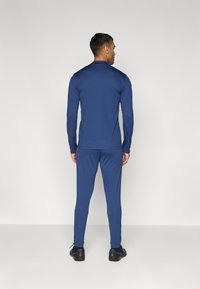 Marineblauw sportoutfit gemaakt van een gladde stof. Langsleeve top en aansluitende broek; gecombineerd met zwarte sportschoenen.