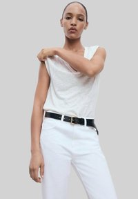 Hvid ærmeløs top med V-hals, parret med hvide jeans og et sort bælte med en guldbuckle. Modellen poserer med den ene arm hævet.