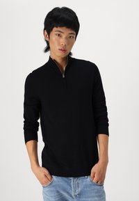 Juodas užtrauktuku užsitraukiantis megztinis su ilgomis rankovėmis, pagamintas iš lygaus, plono audinio. Modelis dėvi šviesiai mėlynas džinsines kelnes.