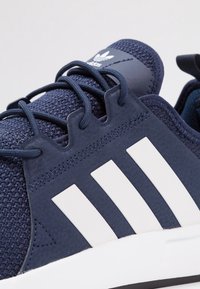 Marinblå atletisk sko med ett texturerat mesh-övre, gummi-accenter, tre vita ränder på sidan och platta snören.