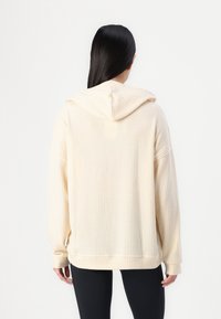 Hellbeige Waffelstrick-Hoodie mit lockerer Passform, Kapuze und langen Ärmeln; strukturiertes Material und gerippte Bündchen. Schwarze Leggings sind darunter sichtbar.
