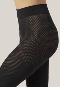 Närbild av en person som bär svarta mönstrade strumpbyxor med hög midja mot en neutral bakgrund.