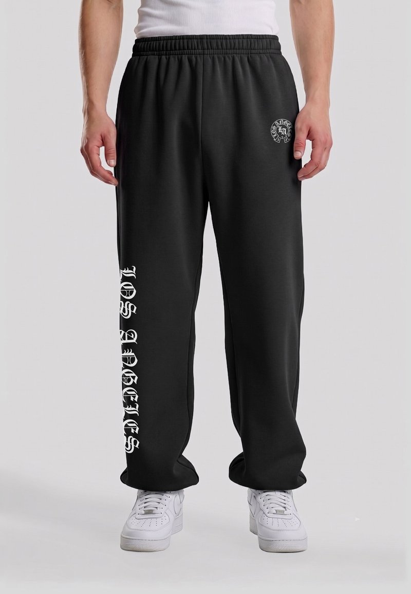 Homme portant un pantalon de survêtement noir avec un texte gothique blanc sur la jambe gauche et un petit logo rond sur la cuisse droite, assorti à des baskets blanches.