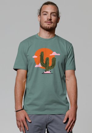 Green cotton t-shirt featuring a cactus graphic in dark green with a pink flower and stylized orange sun and pink clouds. Short sleeves.