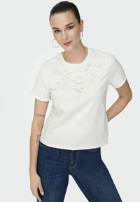 Hvid bomulds t-shirt med et blomster-laserudskæringsdesign på fronten, rund halsudskæring, og korte ærmer, parret med blå jeans.