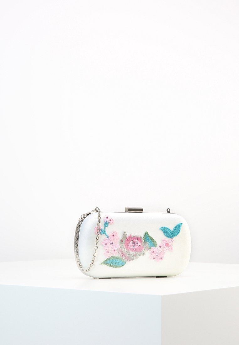 Pochette rectangulaire blanche avec chaîne argentée et broderies florales roses et vertes, présentée sur un piédestal blanc sur fond uni.