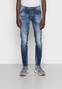 Jeans in denim blu con un design slim fit, caratterizzati da toppe strappate e dettagli con macchie di vernice. Abbinati a sneaker grigie.