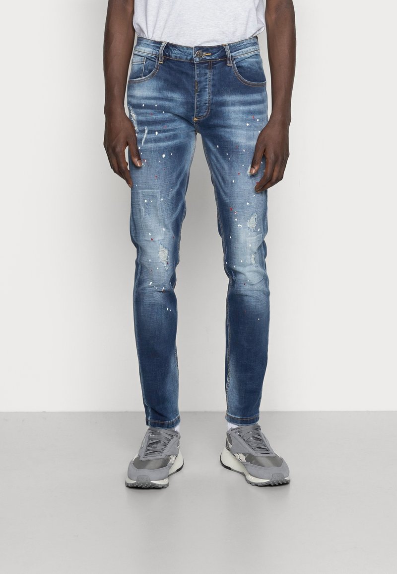 Jeans in denim blu con un design slim fit, caratterizzati da toppe strappate e dettagli con macchie di vernice. Abbinati a sneaker grigie.