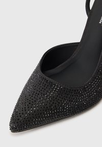 Μαύρες γόβες με μυτερή μύτη που χαρακτηρίζονται από υφή στην επάνω επιφάνεια καλυμμένη με μικρές, γυαλιστερές στρας, με ανοιχτό σχέδιο και λεπτή πίσω αλυσίδα.