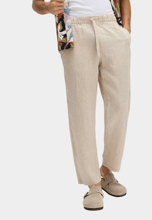 Mand iført beige linnedragter med snøre, slip-on ruskindssko og et mønstret skjorte over en hvid tanktop, stående med hænderne i lommerne.