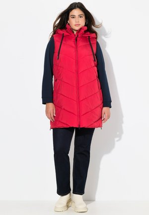 Gilet imbottito rosso con cappuccio, caratterizzato da un design a chevron, cordini regolabili e zip frontale. Indossato sopra una maglia blu navy e jeans scuri.
