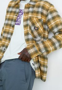 Chemise en flanelle à carreaux jaunes et noirs avec une poche appliquée, portée sur un t-shirt graphique vert clair et un pantalon gris. Tissu texturé.