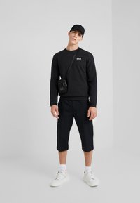 Felpa nera con logo EAZ, pantaloni corti neri e sneakers bianche. Tessuto texturizzato e design dritto. Include una piccola borsa nera.