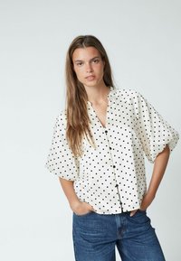 Weiße Bluse mit schwarzen Punkten, kurzen Puffärmeln, Knopfleiste und rundem Kragen, kombiniert mit blauen Jeans.