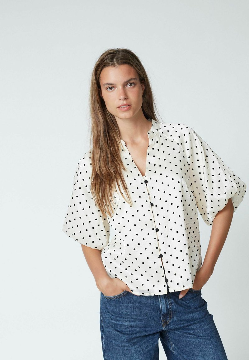 Weiße Bluse mit schwarzen Punkten, kurzen Puffärmeln, Knopfleiste und rundem Kragen, kombiniert mit blauen Jeans.