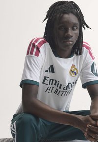 Νέος με dreadlocks φοράει λευκή φανέλα της Ρεάλ Μαδρίτης και πράσινο παντελόνι κάθεται με τα χέρια σταυρωμένα.