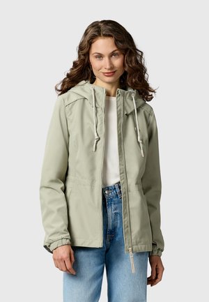 Kobieta o kręconych włosach, ubrana w jasnooliwkową kurtkę z kapturem na białą koszulę i niebieskie dżinsy, stojąca na jasnym tle.