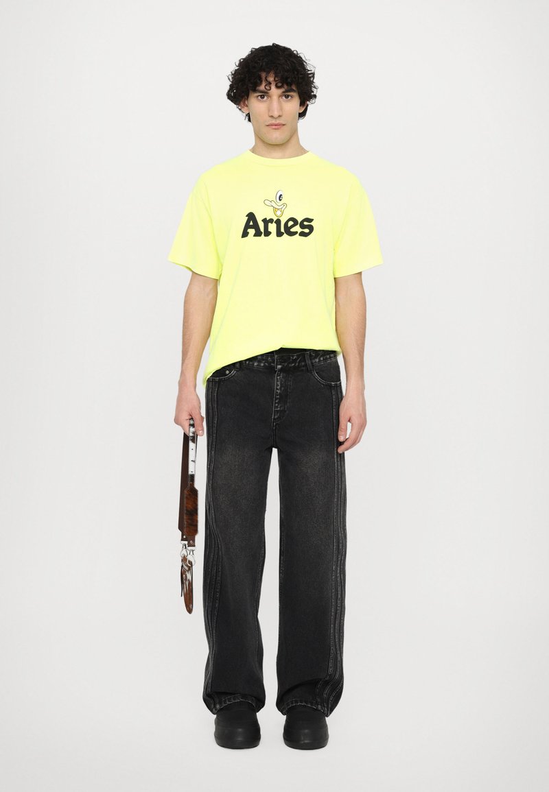 T-shirt gialla oversize con grafica nera "Aries", abbinata a jeans neri a gamba larga con cuciture a contrasto e calzature nere.