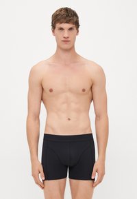 Junger Mann mit kurzen braunen Haaren, der schwarze, eng anliegende Boxer Shorts trägt und vor einem schlichten weißen Hintergrund steht.