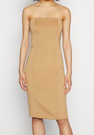 Sukienka beżowa bez ramiączek, do kolan, dopasowana do osoby z widocznymi ramionami i górną częścią tułowia na jednolitym jasnym tle.