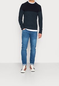 Dunkelblau und marineblauer, farblich blockierter Pullover mit geripptem Muster, kombiniert mit hellblauen Jeans und weißen Sneakers mit braunen Akzenten.