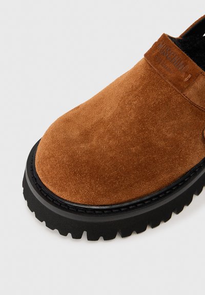 Chaussure slip-on en daim marron avec une épaisse semelle en caoutchouc noir, présentant une finition texturée et un marquage discret sur le côté.
