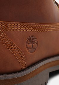 Stivale in pelle marrone con finitura testurizzata, caratterizzato da cuciture dettagliate e un logo ad albero embossato sul lato. Suola in gomma resistente visibile.