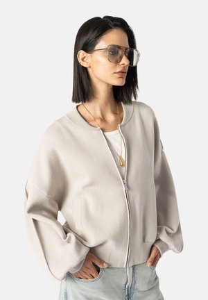 Jasno beżowa kurtka zapinana na zamek z szerokimi rękawami w kształcie dzwonu, o miękkiej fakturze i dwóch przednich kieszeniach. Połączona z białą bluzką i jasnoniebieskimi dżinsami.