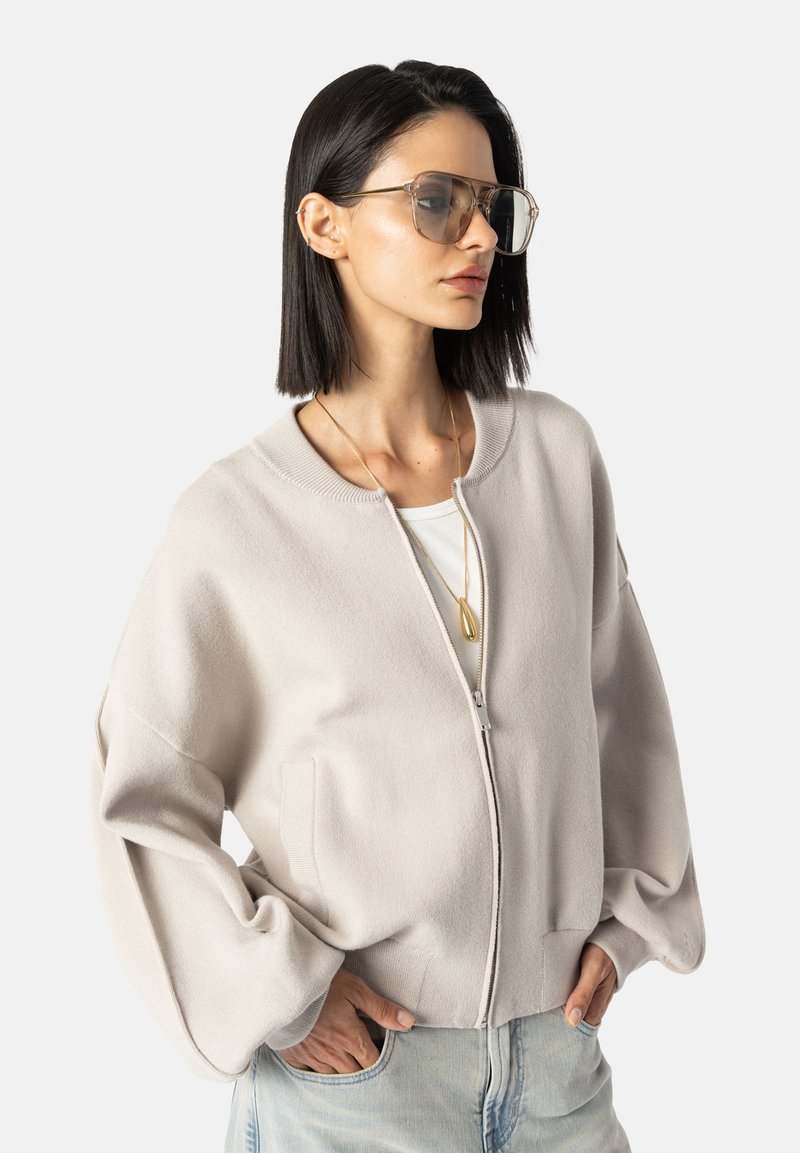 Chaqueta beige claro con cremallera, mangas acampanadas amplias, textura suave y dos bolsillos frontales. Combinada con una blusa blanca y jeans azul claro.