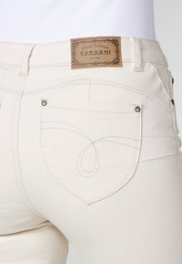 Jeans de mezclilla color crema con una etiqueta de cuero que dice "KOROSHI" y costuras decorativas en los bolsillos traseros. Textura suave, diseño ajustado.