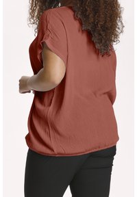 Rostfarbene Bluse aus strukturiertem Stoff, mit kurzen Ärmeln und lockerer Passform, kombiniert mit schwarzen Hosen. Verfügt über einen dezenten gerafften Saum.