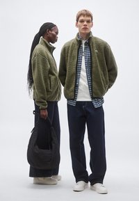 Olivengrønne fleecejakker med høje kraver og lynlåslommer, båret med mørk denim. Sort quiltet taske og hvide sneakers fuldender looket.