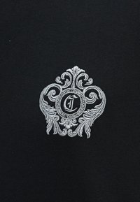 Περιτεχνή λευκή κεντητική σε μαύρο ύφασμα με κεντρικό κυκλικό έμβλημα που φέρει το γράμμα "J" και διακοσμητικά σχέδια γύρω του.