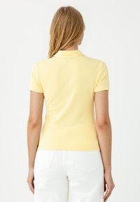 Polo shirt giallo a costine con maniche corte, dotato di colletto. La maglietta è aderente, abbinata a pantaloni bianchi, e presenta una trama liscia.