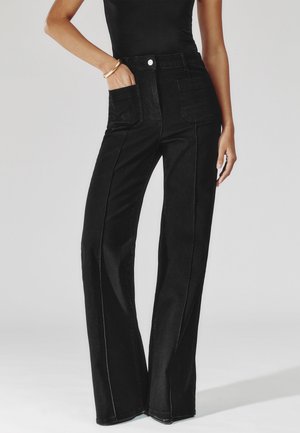 Donna che indossa pantaloni neri a vita alta e gambe larghe con tasche applicate sul davanti, abbinati a un top nero senza maniche e braccialetti dorati.