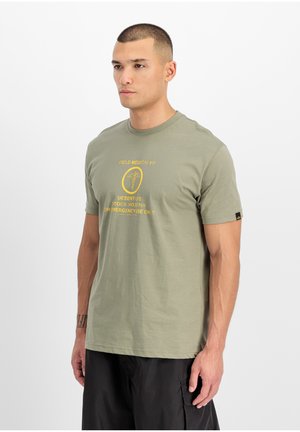 Man wearing a gray-green T-shirt with yellow "Field Medical Kit" text and black cargo shorts, standing against a plain white background.