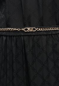 Λεπτομέρεια μαύρου φορέματος με υφή υφάσματος και διακριτικό σχέδιο. Μια χρυσή αλυσίδα ζώνης με έντονο λογότυπο τονίζει τη μέση.