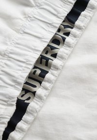 Tessuto bianco con texture arricciata, presenta un'etichetta blu navy e crema con la scritta "SUPERDRY" lungo il bordo. Finitura liscia e leggero lucido.