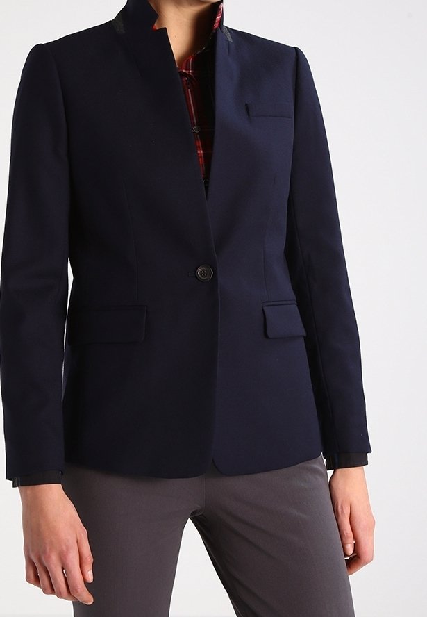 Blazer bleu marine avec col montant, fermeture par un bouton unique, deux poches avant, et un léger brillant. Superposé à une chemise à carreaux.